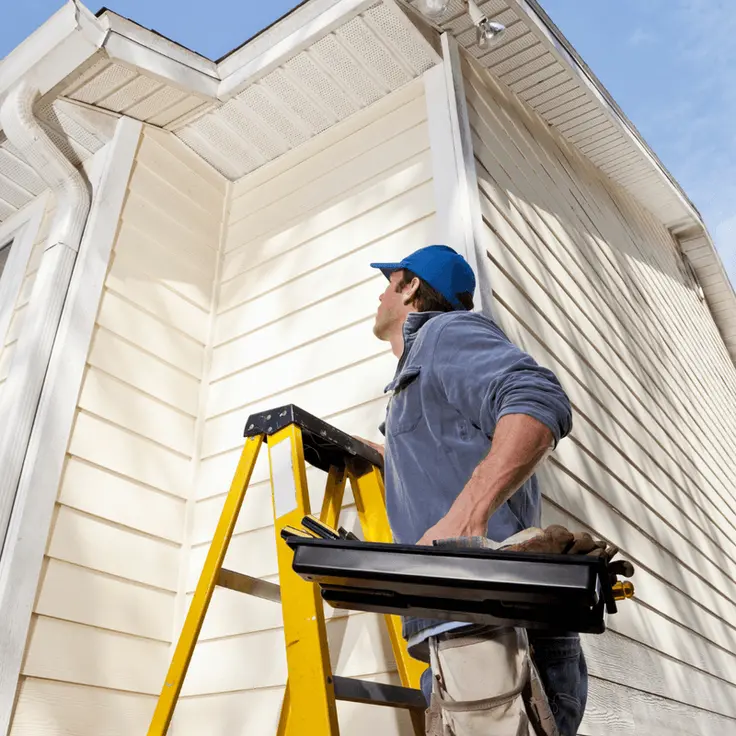 Worker wearing blue cap and jacket climbing yellow ladder outside a beige house, carrying a toolbox.