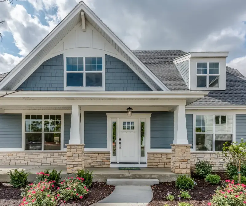 The front exterior of a blue craftsman-style home with a covered porch, stone column bases, and landscaped flower beds