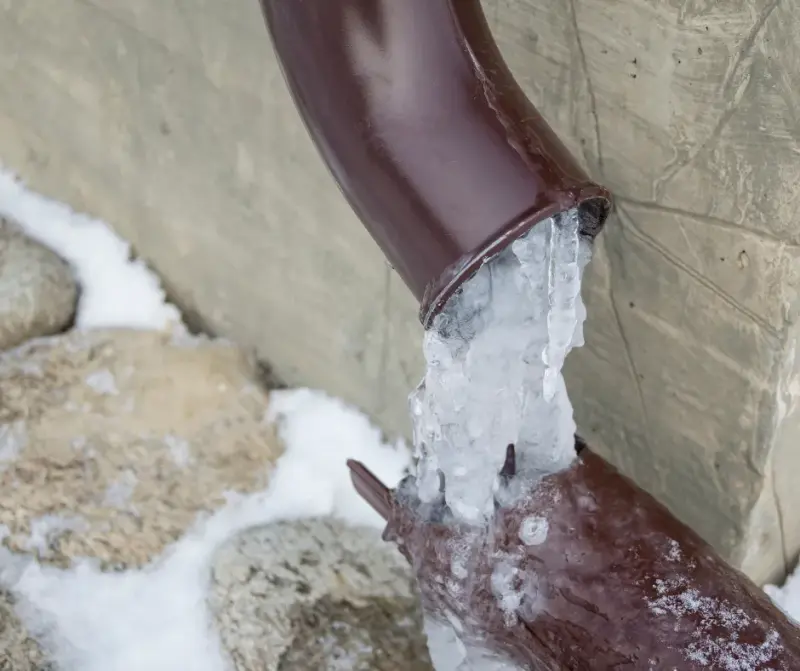 A downspout clogged with ice, with frozen water spilling over its edge against a snowy stone foundation