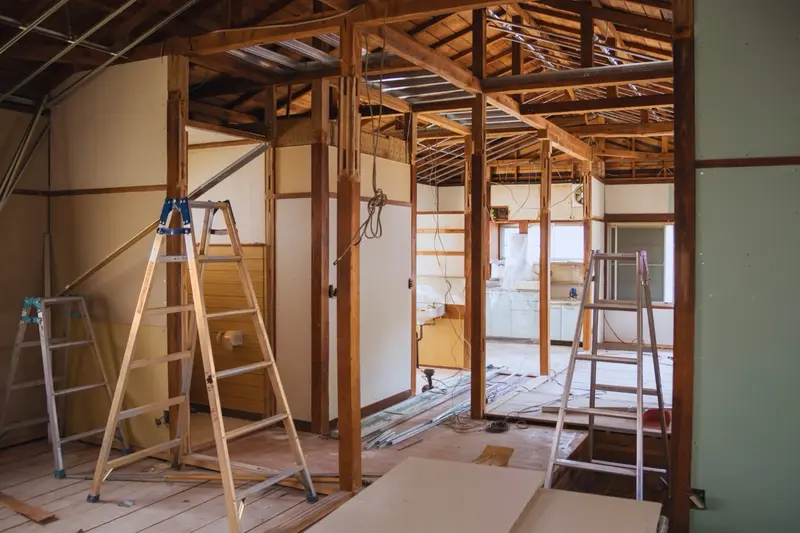 A home interior stripped to its wooden framing during a major renovation, with ladders and drywall panels staged across the open floor