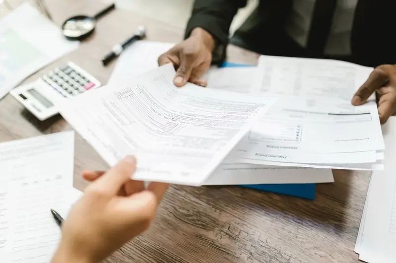 Two people exchanging tax forms and financial statements across a desk with a calculator nearby