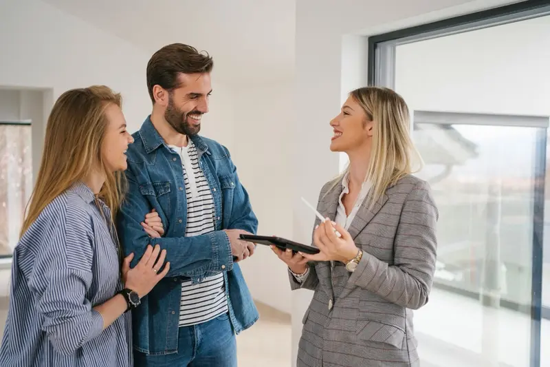 A smiling couple reviewing paperwork on a tablet with a real estate agent inside a bright, empty home